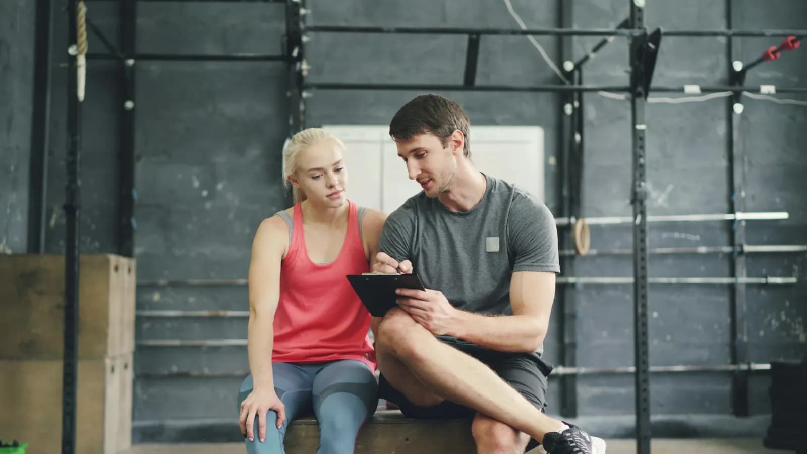 A trainer going over a client’s plan on a tablet, both sitting on a gym bench.