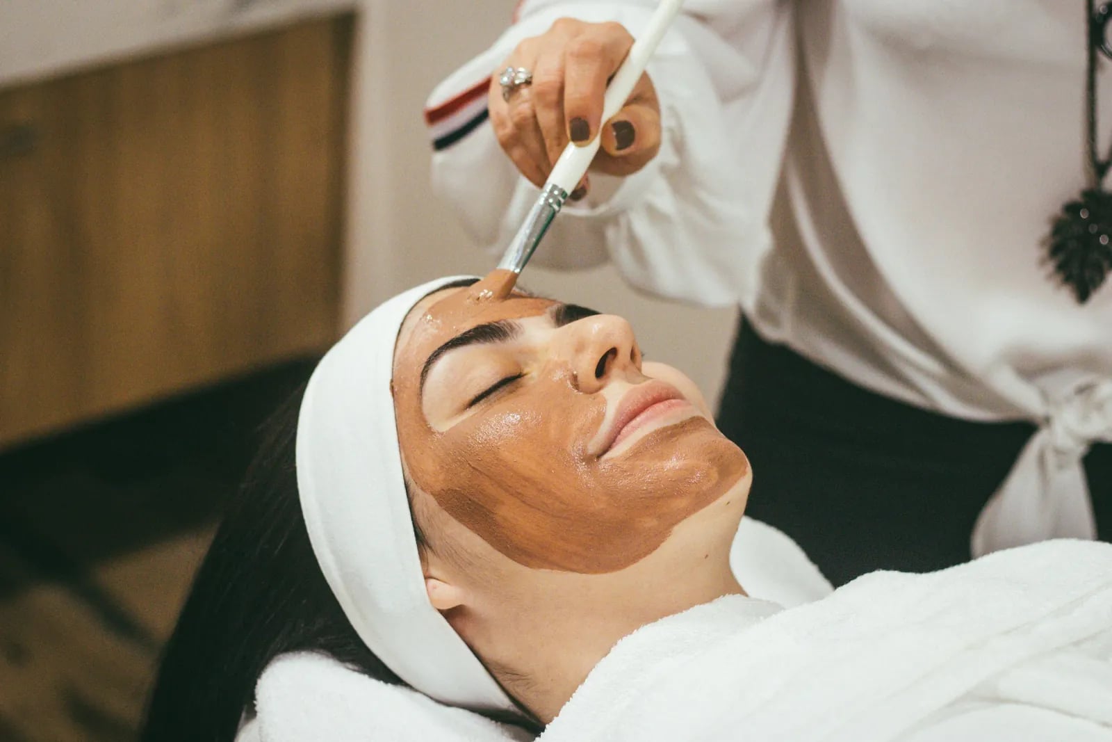 An esthetician brushing a clay mask onto a relaxed client’s face.